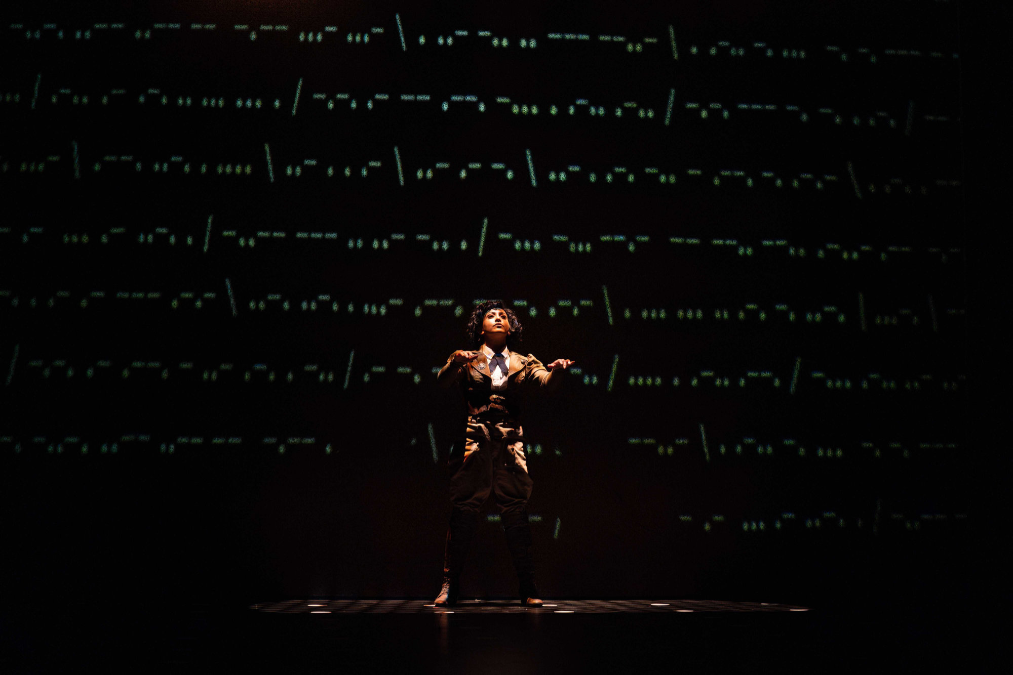 A woman stands centre stage, posed as if in the midst of coding. The backdrop features lines of dots and dashes.