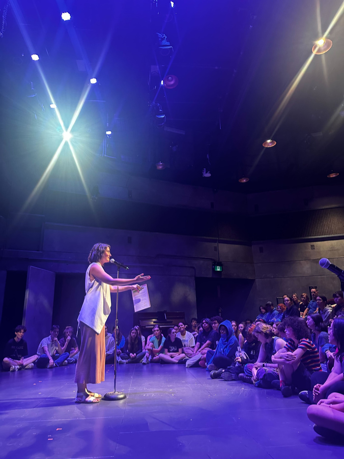 A woman in a white top and brown skirt stands at a microphone, addressing a group of students who sit facing her on stage.