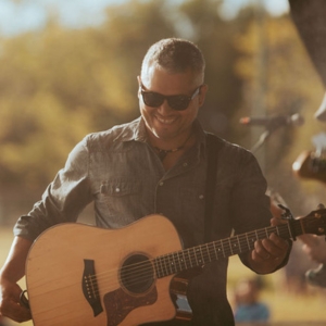 Don Amero smiles as he stands on an outdoor stage with his guitar.