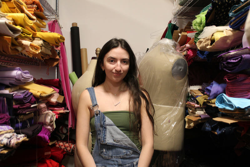 A young woman wearing a green top and overalls stands between two tall shelves of colourful fabric in the wardrobe department.