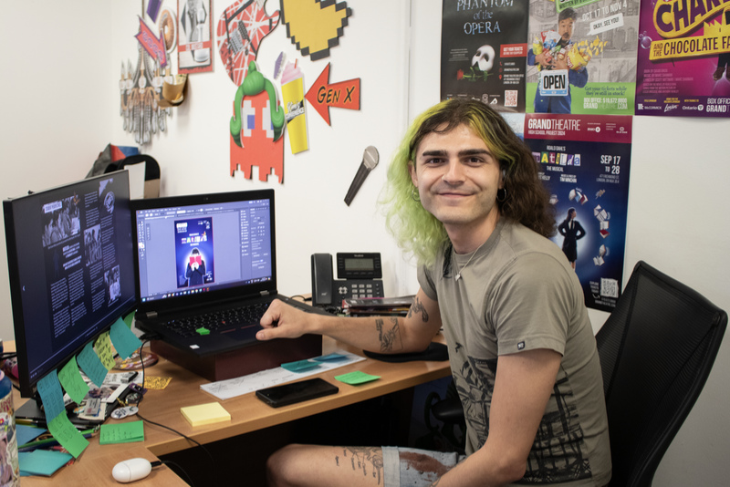 A young adult wearing a brown top and denim shorts smiles as they sit at their desk in the Grand Theatre's Marketing department, surrounded by show posters and colourful post-it notes.