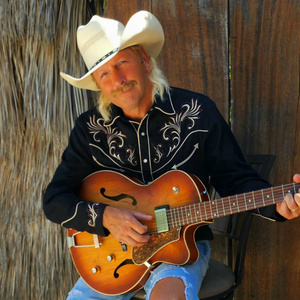 A musician, Aaron Halliday, wears a black western shirt, jeans, and white cowboy hat as he poses with his guitar. A macaw is superimposed on the right side of the photo.