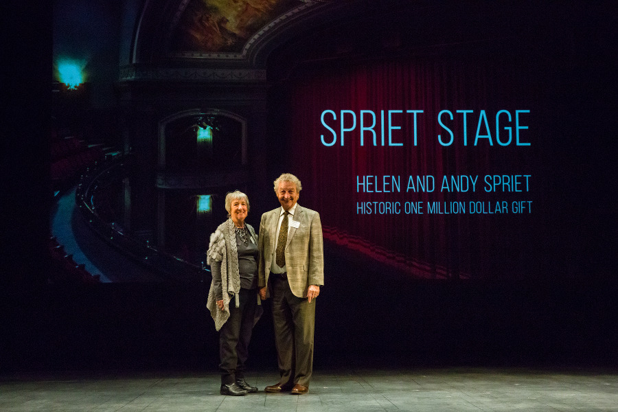 Helen and Andy Spriet stand smile as they stand on the Spriet Stage. Projected behind them is a photograph of the Stage from the balcony level, featuring an angled view of the proscenium arch.