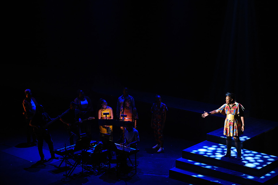Alexandra Kane stands in the spotlight on stage during a performance of Finding Black Joy. To her left are several musicians and singers.