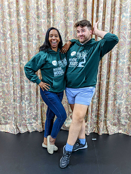 Saccha and Greg pose in front of a curtain in the rehearsal hall, wearing matching Into the Woods hooded sweatshirts.
