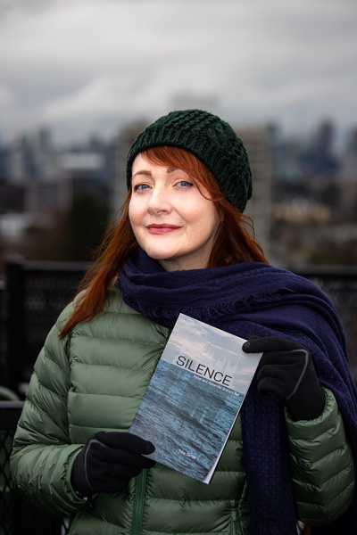 Trina Davies stands outdoors, wearing a winter coat, hat and scarf. She holds a copy of her play 'Silence: Mabel and Alexander Graham Bell'.