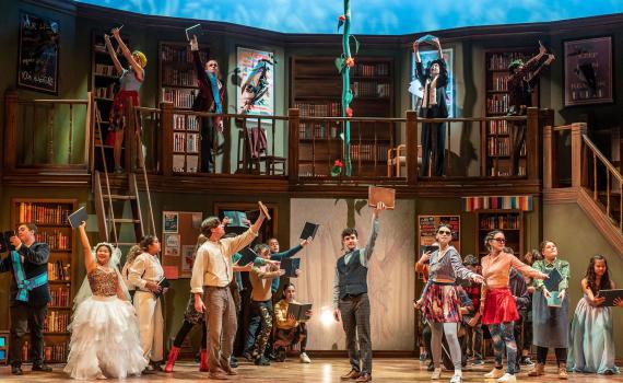 The cast of Into the Woods stands staggered on the Spriet Stage holding books triumphantly in the air. The background set emulates a library and a large plant bean stock grows in the back middle. Photo by Dahlia Katz.
