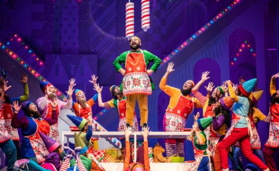 Members of the cast of ELF (18 people) stand in a group formation around the character of Buddy the Elf. Buddy wears a green and yellow elf costume and red holiday apron, and stands high on a table. Surrounding cast members wear a variety of colourful, holiday apparel – along with fake beards – to appear as elves of the North Pole. In the background, Christmas lights and a sparkly, Christmas-themed set are visible. Photo by Dahlia Katz