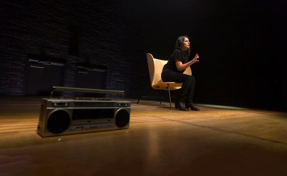 Zorana Sadiq wears all black and sits on a large wooden chair on a mostly bare stage. She leans forward, and her hands are being used expressively to convey a point. In the foreground lies a silver boom box.
