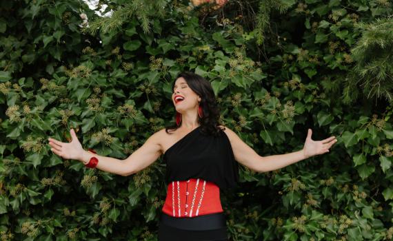 Andrea Menard stands in front of some greenery, wearing a black and red dress with her arms outstretched.