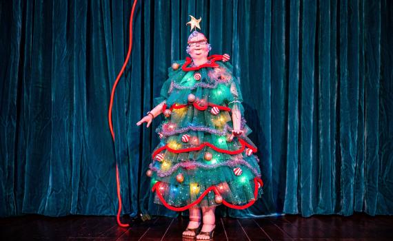 A marionette in a dress that resembles and is decorated like a christmas tree stands in front of a teal curtain.