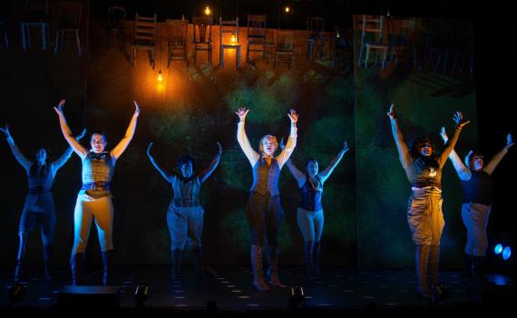The cast of The Invisible stands on stage with arms in the air, warm lighting downstage shifts to a cool blue lighting upstage. A row of wooden chairs hangs high above the cast against the backdrop.