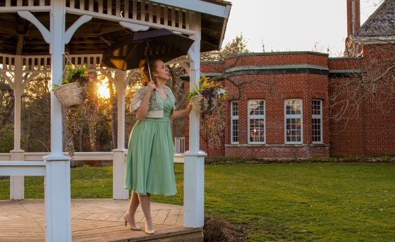 A woman wearing a green dress holds an umbrella as she stands at the edge of a gazebo.