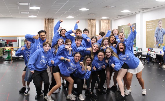 A group of teens in matching light blue Matilda hoodies lean in to pose for a photo in the rehearsal hall.