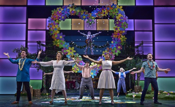 The cast of As You Like It stands in the foreground with outspread arms. A large heart made up of colourful flowers frames another performer with a guitar as they stand on a platform in the background.