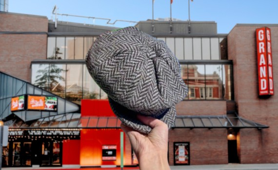 A newsboy cap is held up in the foreground, the Grand Theatre visible in the background.