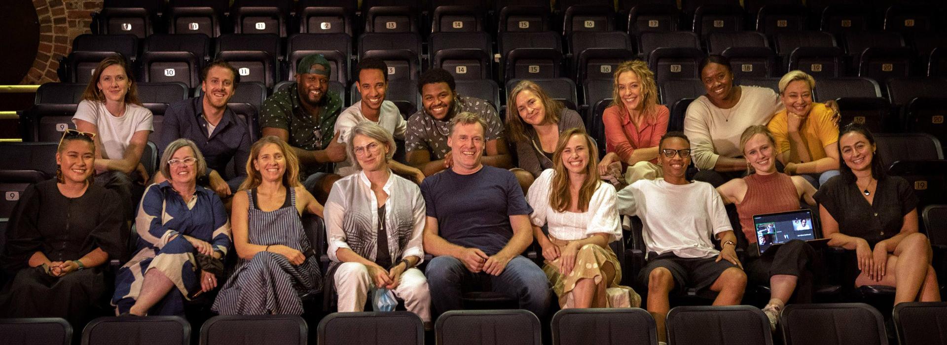 The company of Fall On Your Knees sits in two rows of an intimate theatre space and smiles at the camera. The theatre chairs are dark in colour and the back wall is a multi-shade brick. Photo by John Lauener Photography