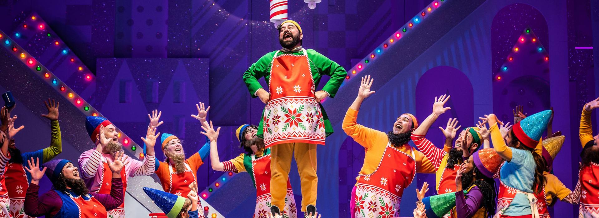 Members of the cast of ELF (18 people) stand in a group formation around the character of Buddy the Elf. Buddy wears a green and yellow elf costume and red holiday apron, and stands high on a table. Surrounding cast members wear a variety of colourful, holiday apparel – along with fake beards – to appear as elves of the North Pole. In the background, Christmas lights and a sparkly, Christmas-themed set are visible. Photo by Dahlia Katz