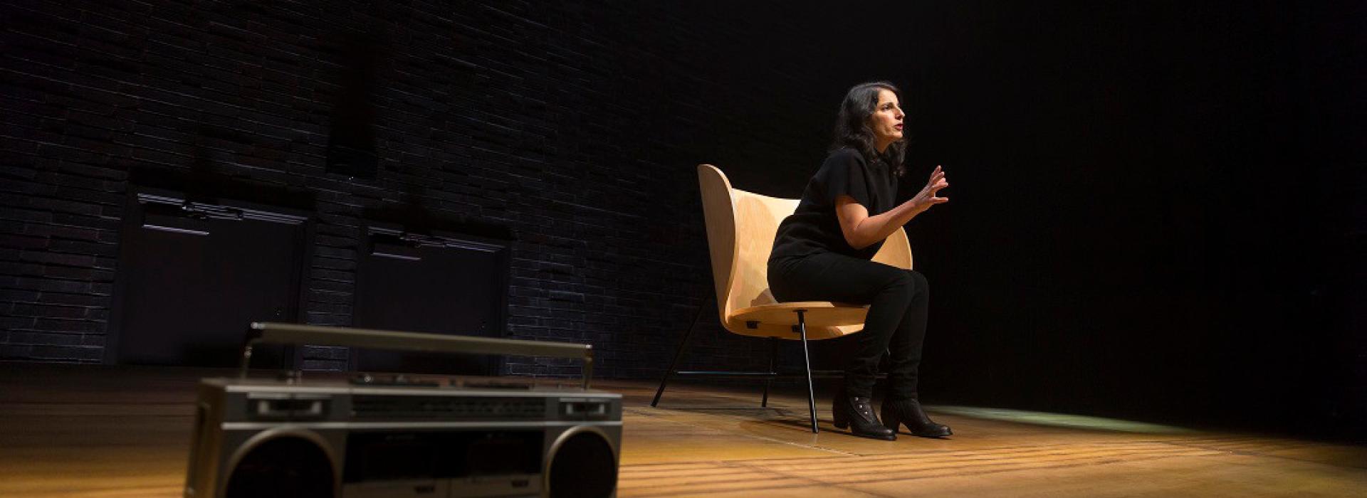 Zorana Sadiq wears all black and sits on a large wooden chair on a mostly bare stage. She leans forward, and her hands are being used expressively to convey a point. In the foreground lies a silver boom box.