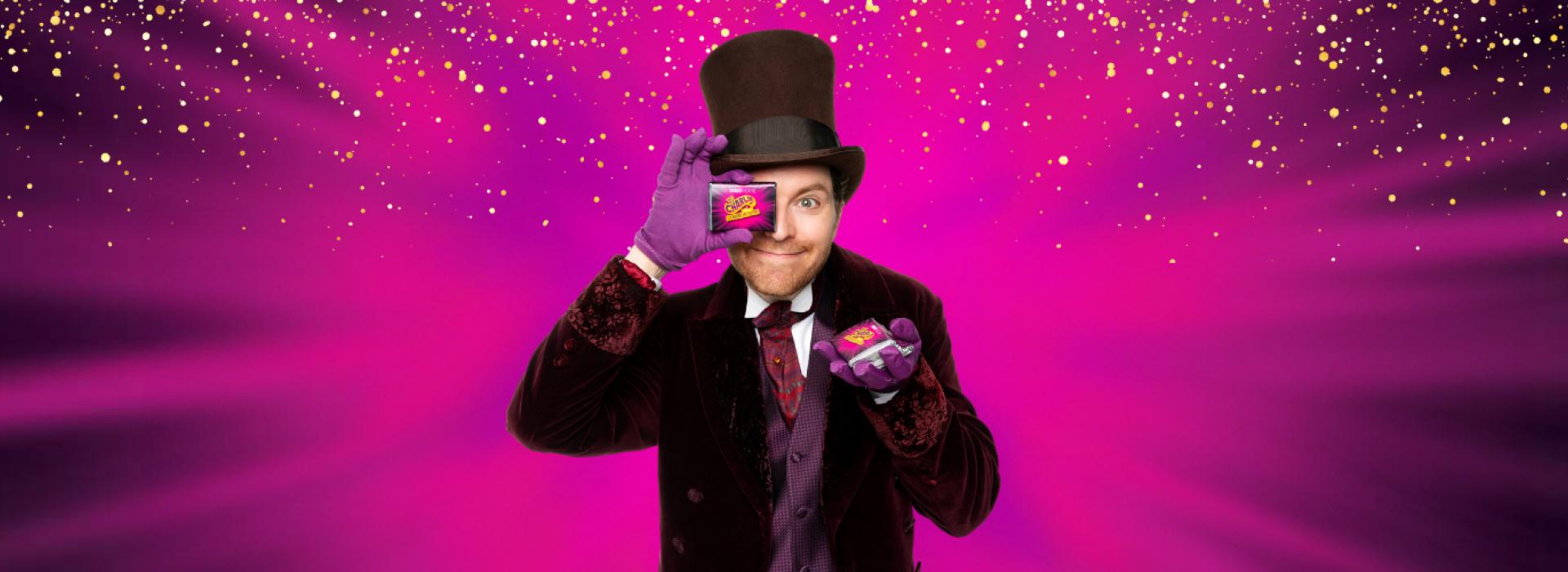 Mark Uhre as Willy Wonka holds up a chocolate bar, in front of a vibrant pink and purple background.