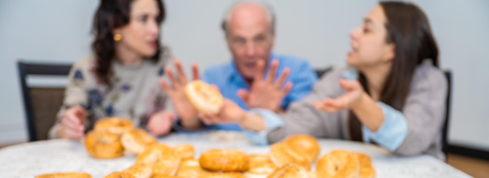 Cast of In Seven Days sits at a dinner table, a pile of bagels in focus in the foreground.