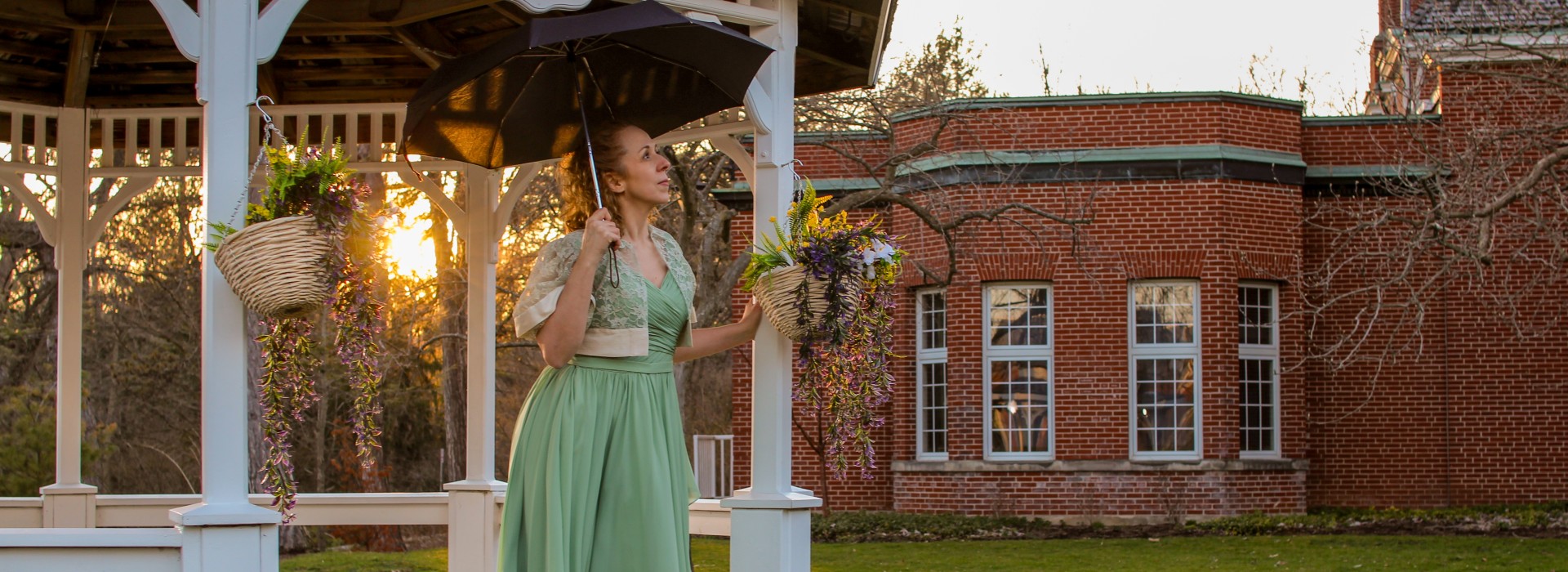 A woman wearing a green dress holds an umbrella as she stands at the edge of a gazebo.