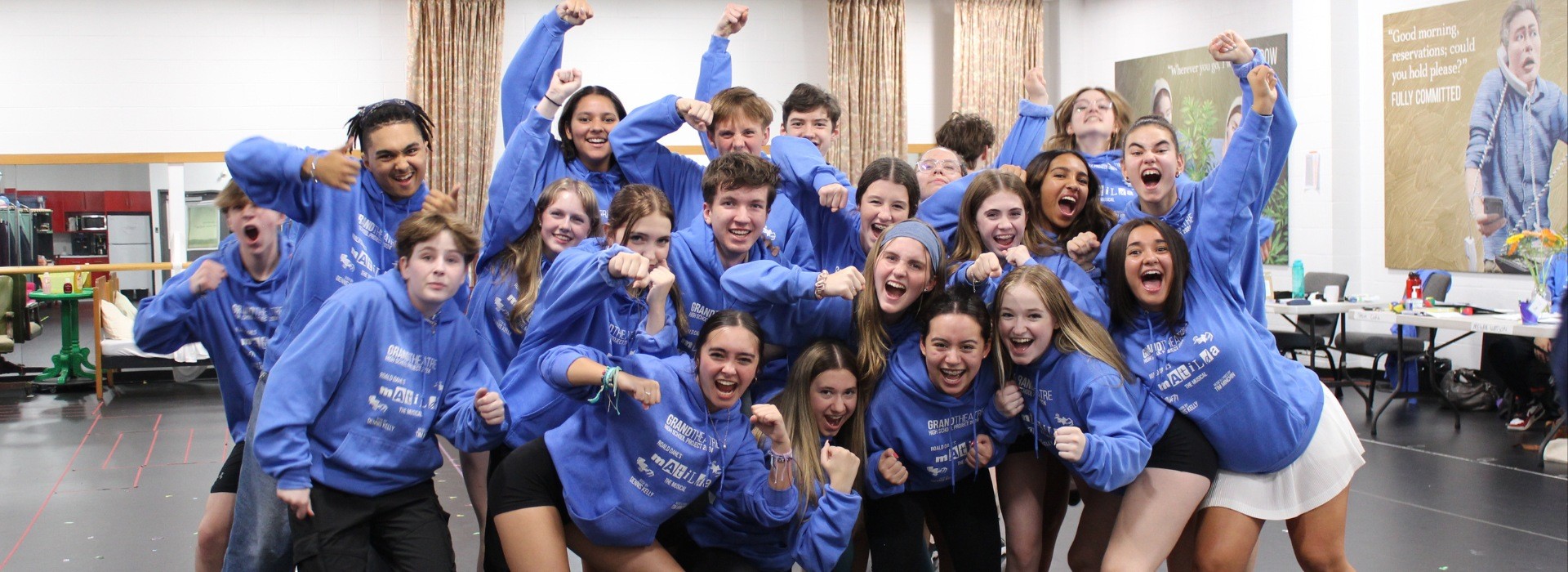 A group of teens in matching light blue Matilda hoodies lean in to pose for a photo in the rehearsal hall.