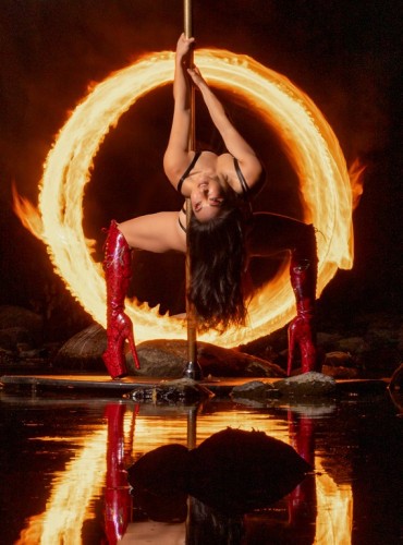 A woman poses while poledancing, a ring of fire in the background.