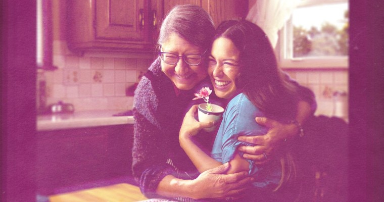 An older woman hugs a younger woman seated at a kitchen table as they enjoy a cup of tea.