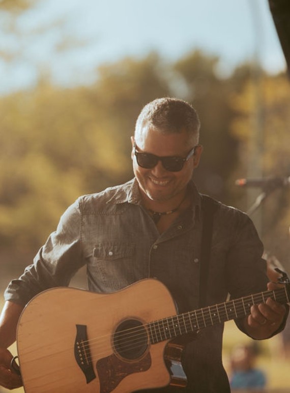 Don Amero smiles as he stands on an outdoor stage with his guitar.